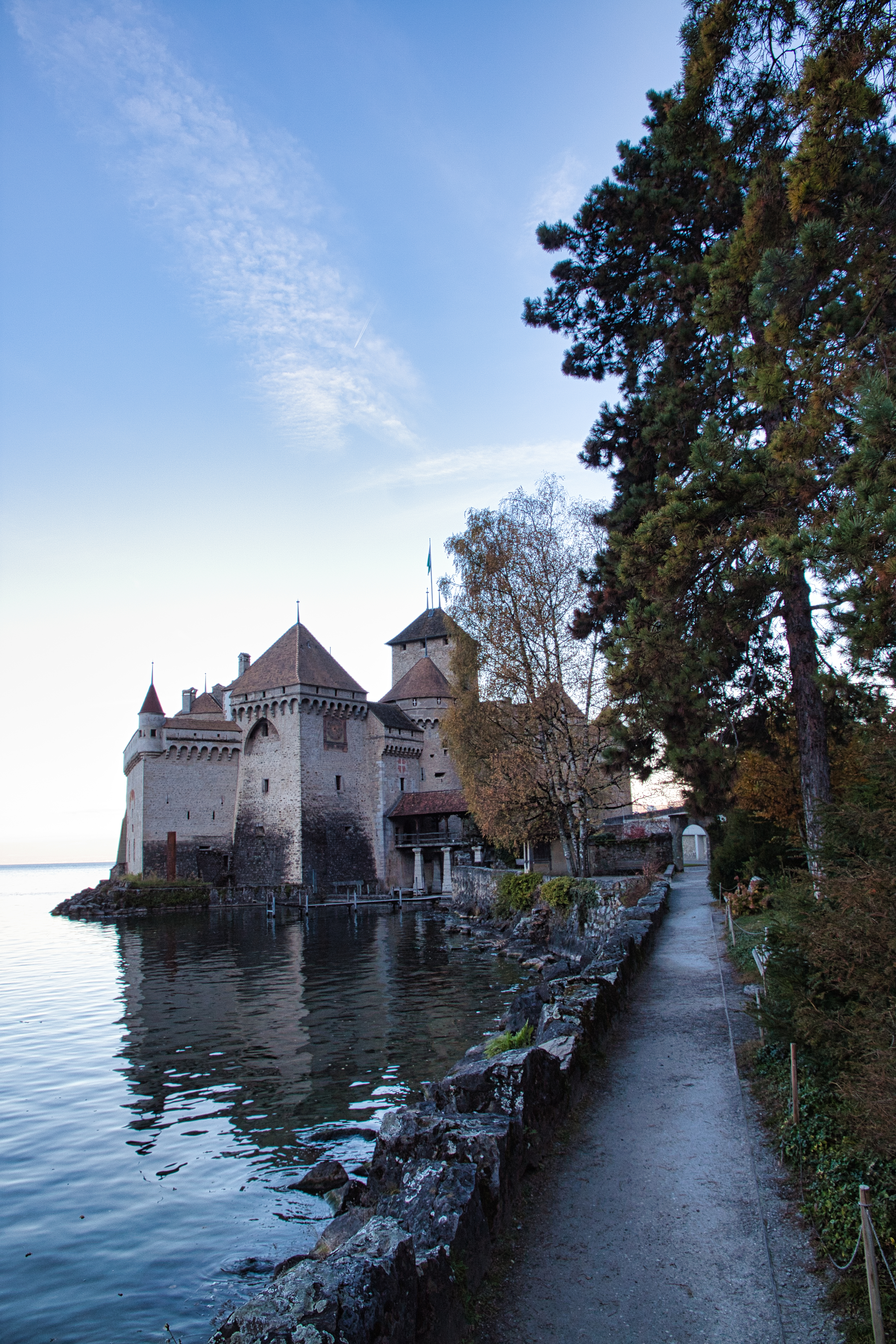 Le château de Chillon vu du sud : ses tours et son corps de logis, couverts de toits de tuiles, se dressent sur un rocher au bord du Léman, longé par un chemin de rive bordé d'arbres.