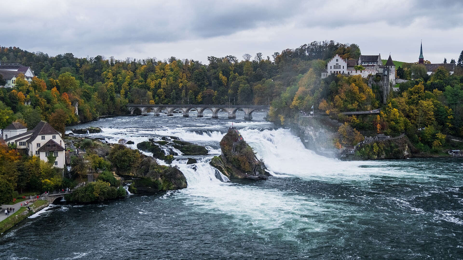 Les chutes du Rhin en pleine largeur : la nappe d'eau blanche se divise autour du rocher central couvert de végétation ; sur la rive dominant les chutes se dresse le château de Laufen, entouré d'arbres aux couleurs d'automne, et un pont enjambe le fleuve en amont.
