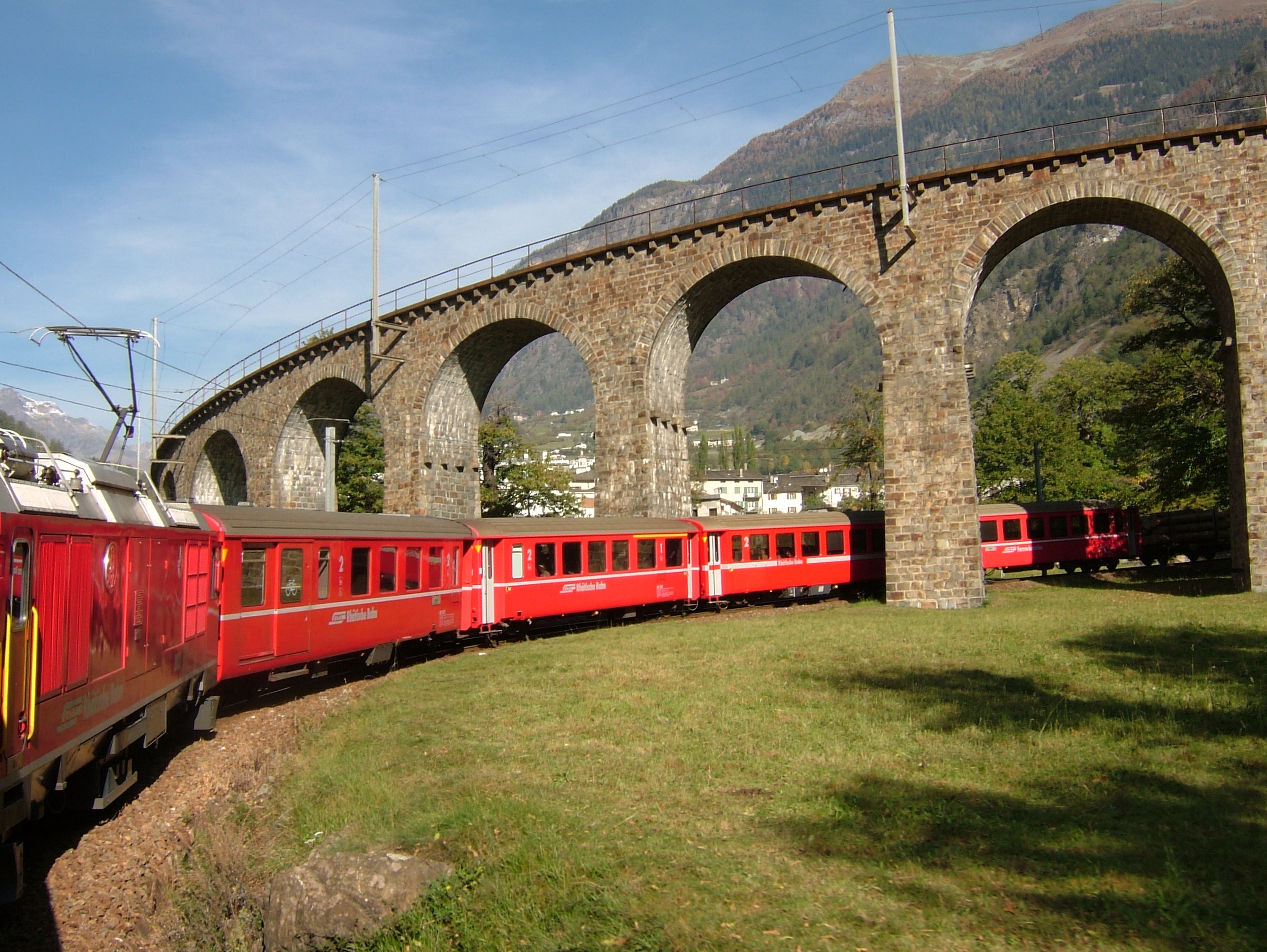 Un train rouge des Chemins de fer rhétiques longe en courbe le viaduc hélicoïdal de Brusio, dont les arches de pierre portent la voie à l'étage supérieur de la boucle.