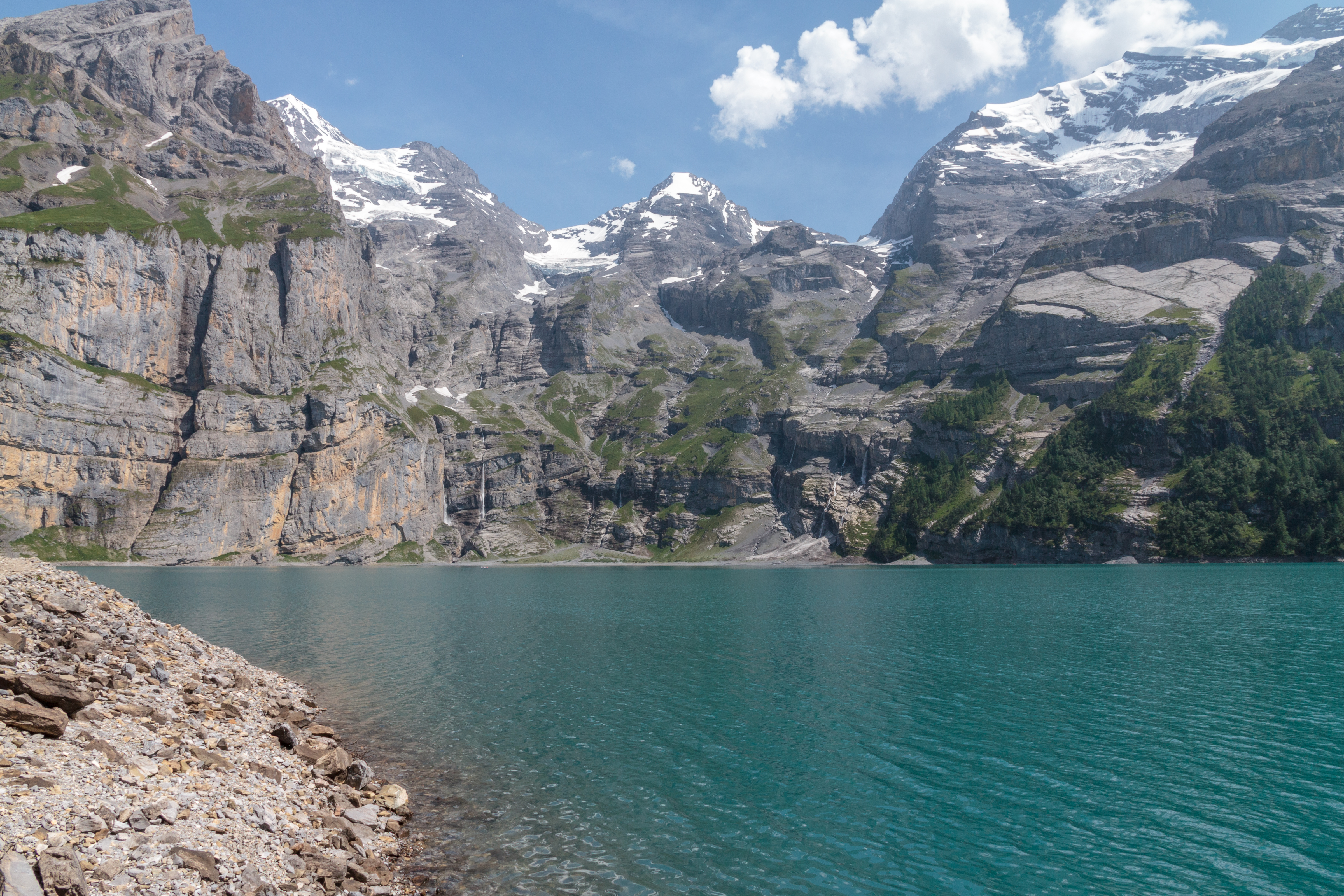 Les eaux turquoise du lac d'Oeschinen au pied de hautes parois calcaires striées de névés ; une grève de galets occupe le premier plan.